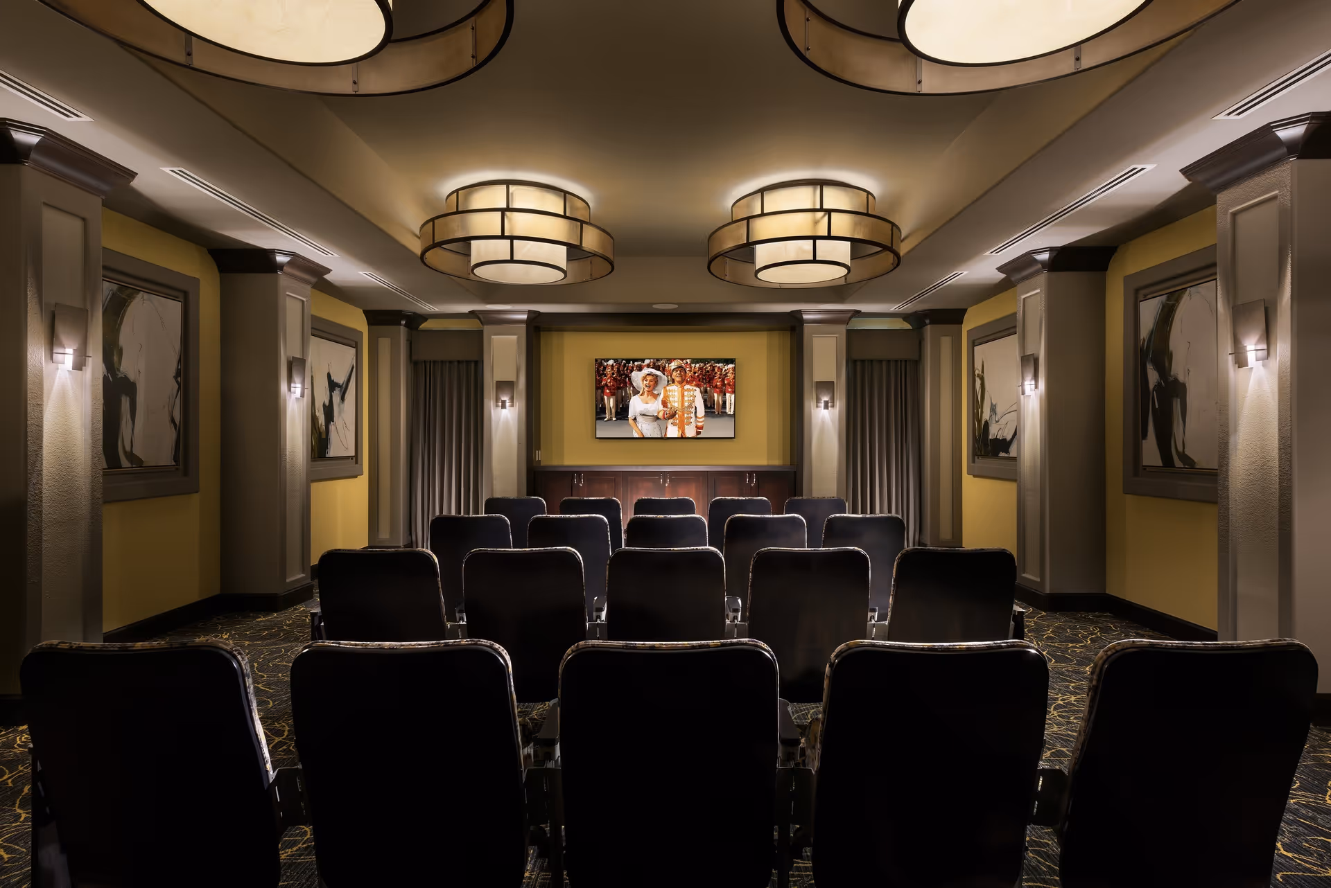 A small theater room with three rows of black cushioned chairs facing a wall-mounted flat screen TV. The room has yellow walls with framed abstract artwork, decorative columns, and three large circular ceiling lights. The carpet has a dark pattern, and the room is softly lit with wall sconces.