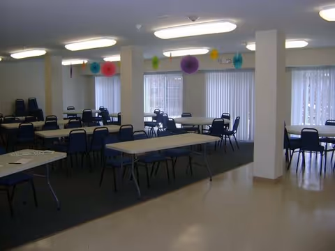 Large multipurpose dining/activity room with folding tables, blue chairs, and colorful paper decorations hanging from the ceiling.