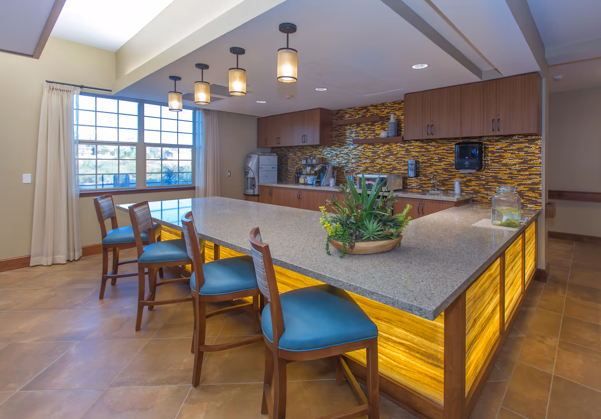 A modern kitchen area with a large granite countertop island surrounded by wooden chairs with blue cushions. The island features under-counter lighting with a warm yellow glow. The back wall has wooden cabinets and a mosaic tile backsplash in shades of brown and yellow. There are pendant lights hanging from the ceiling above the island, a large window with cream curtains letting in natural light, and a decorative plant centerpiece on the island.