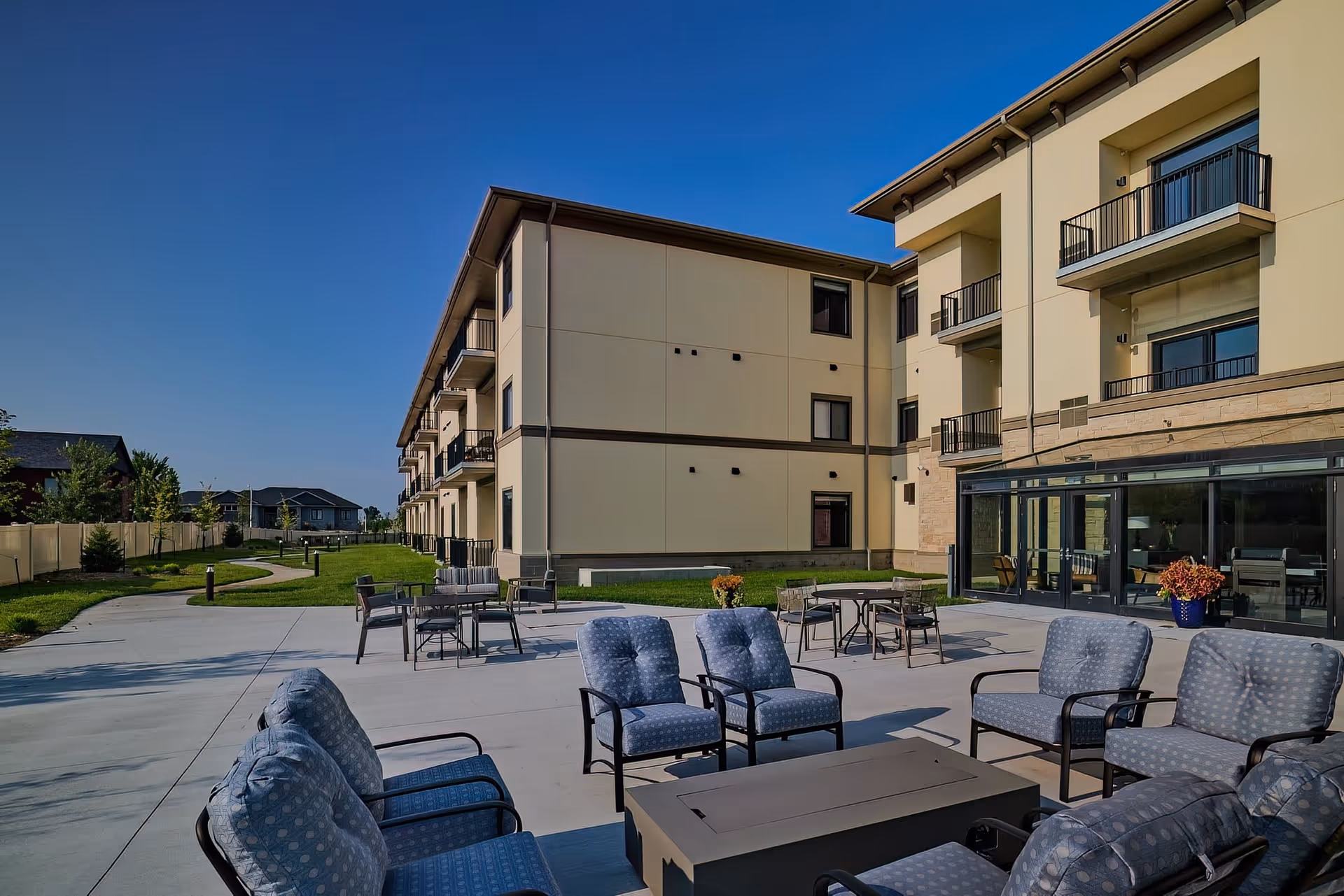 Outdoor patio area at The Parkwood with cushioned chairs arranged around a rectangular fire pit table, additional tables and chairs in the background, and a multi-story building with balconies under a clear blue sky.