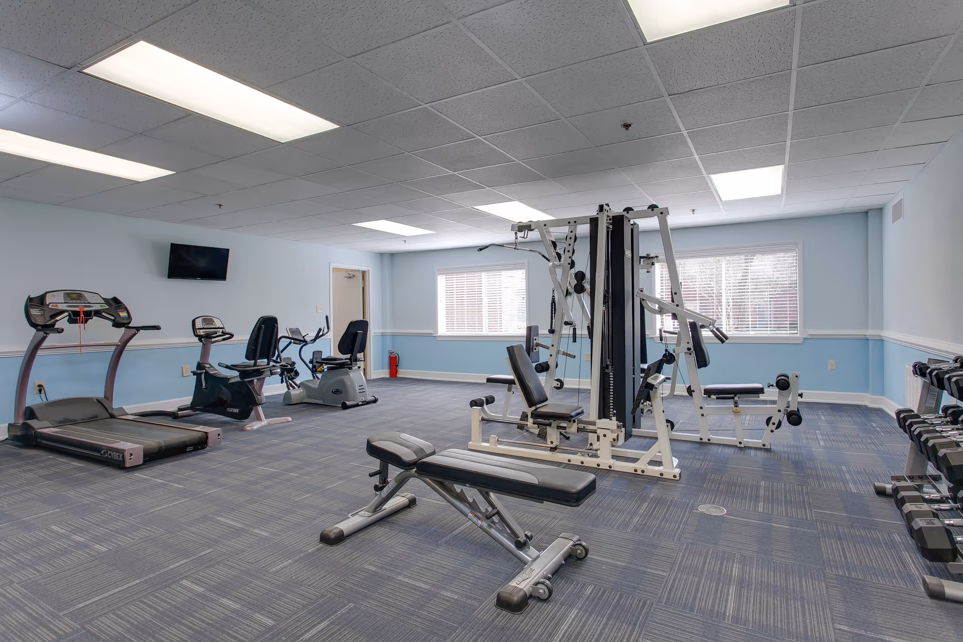 A fitness room with various exercise equipment including a treadmill, stationary bikes, a multi-station weight machine, a bench, and a rack of dumbbells. The room has light blue walls, large windows with blinds, and a wall-mounted TV.