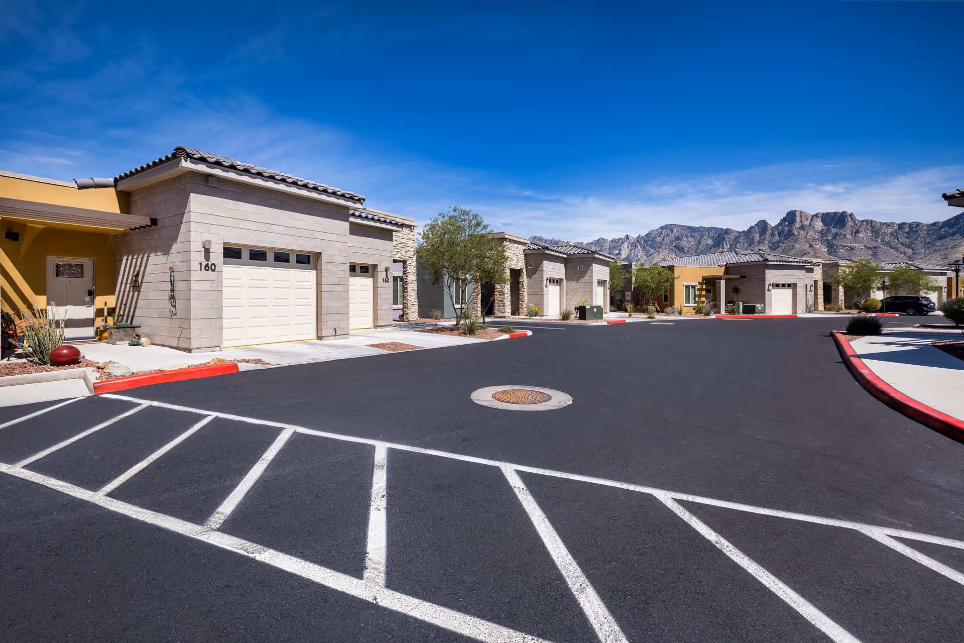 View of a senior living community street with single-story buildings featuring garages and desert landscaping, set against a backdrop of mountains under a clear blue sky.