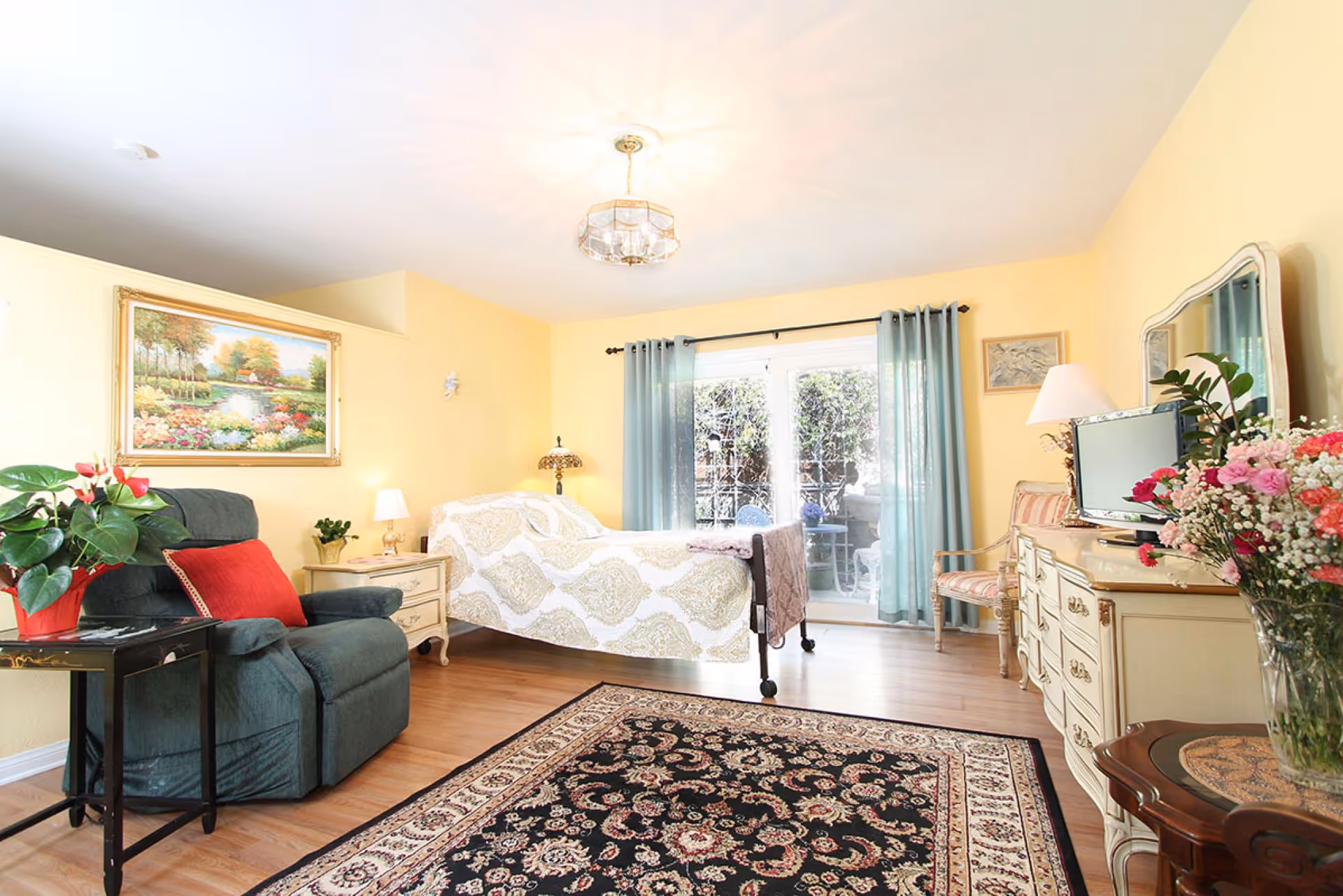 A bright and cozy bedroom with yellow walls, a patterned bed with wheels, a green recliner chair with a red pillow, a floral painting on the wall, a dresser with a mirror and a TV, a side table with a lamp, and a large window with blue curtains leading to an outdoor patio. A decorative rug covers part of the wooden floor.