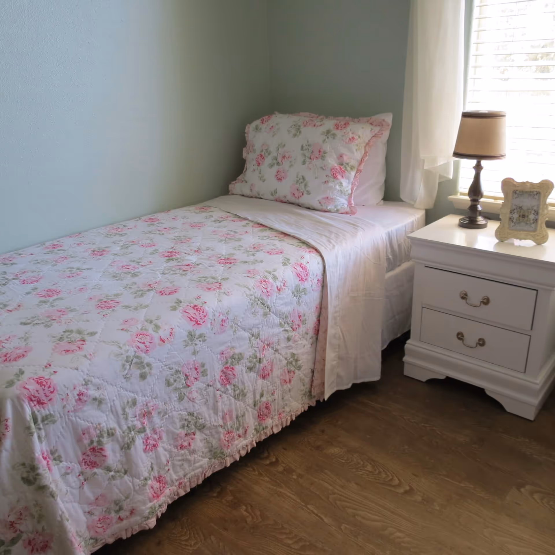 A single bed with a floral patterned quilt and matching pillow in soft pink and green tones, next to a white nightstand with two drawers. On the nightstand, there is a small lamp with a beige shade and a decorative picture frame. A window with white blinds and a sheer curtain is behind the nightstand, allowing natural light into the room.