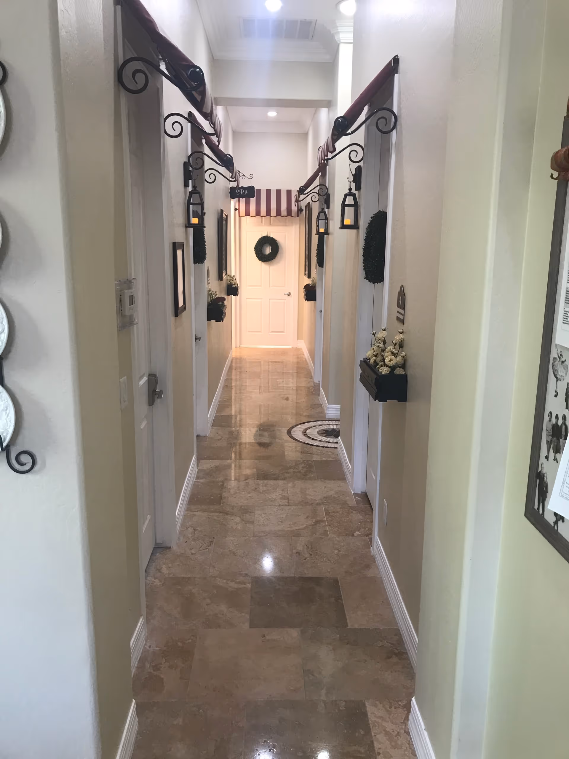 A narrow, well-lit hallway with polished tile flooring and cream-colored walls. The hallway features decorative elements such as small hanging lanterns, flower boxes with white flowers, and green wreaths on the doors. There is a small sign labeled 'SPA' hanging from the ceiling near the end of the hallway, which is closed off by a white door with a green wreath and a striped awning above it.