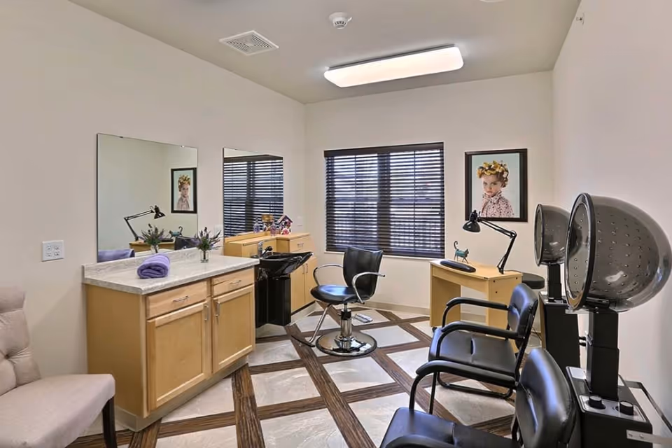 A well-lit hair salon room with two black salon chairs, two hair dryers, a black wash basin, a wooden cabinet with a countertop, two large mirrors, a small desk with a lamp and decorative items, a beige chair, and a framed portrait on the wall. The room has a window with blinds and a patterned floor.
