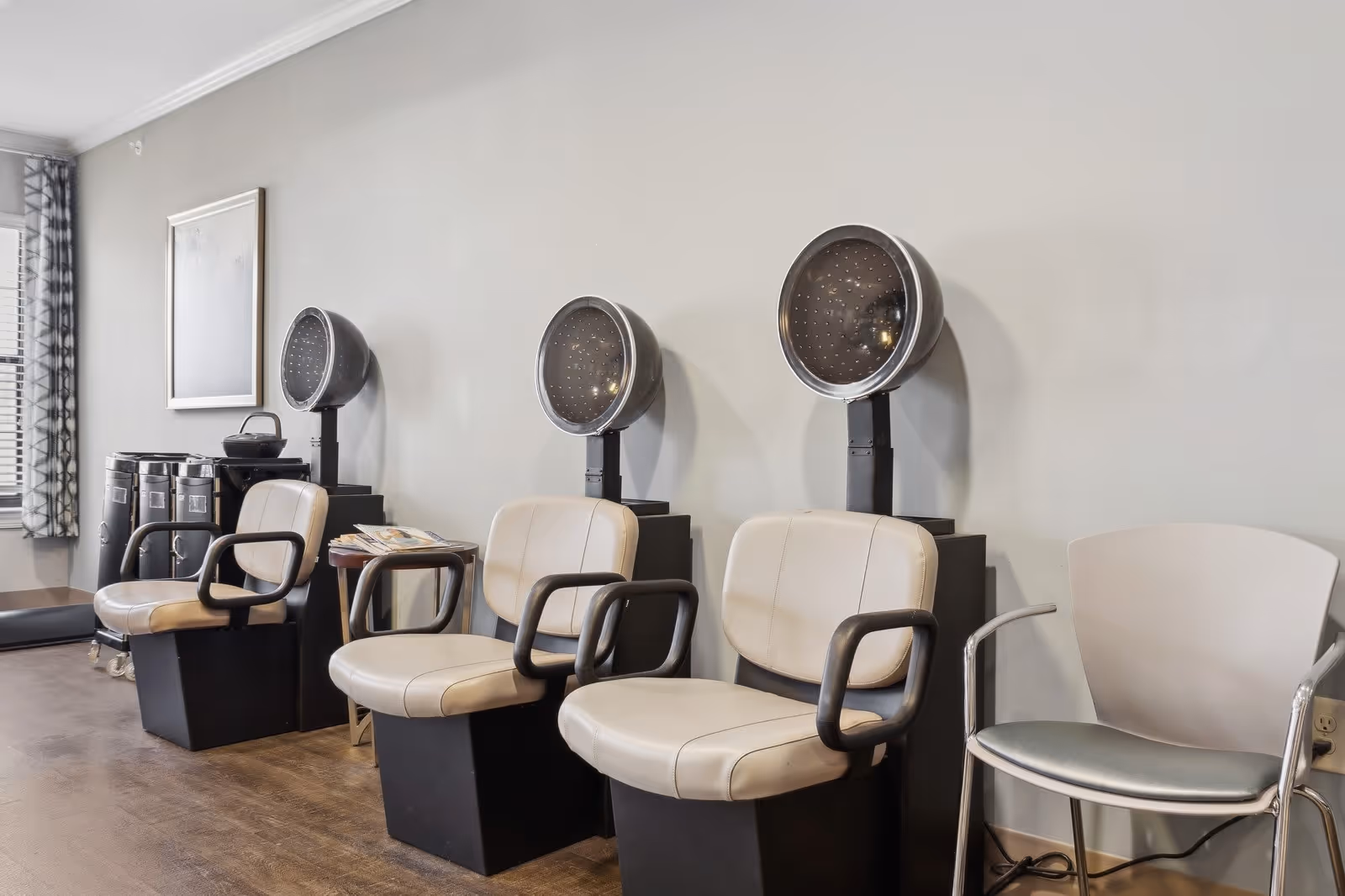 Interior view of a hair salon area with three beige salon chairs each positioned in front of a black hair dryer hood. There is a small table with magazines between two of the chairs, a window with patterned curtains on the left, and an additional gray chair on the right side against a light gray wall.