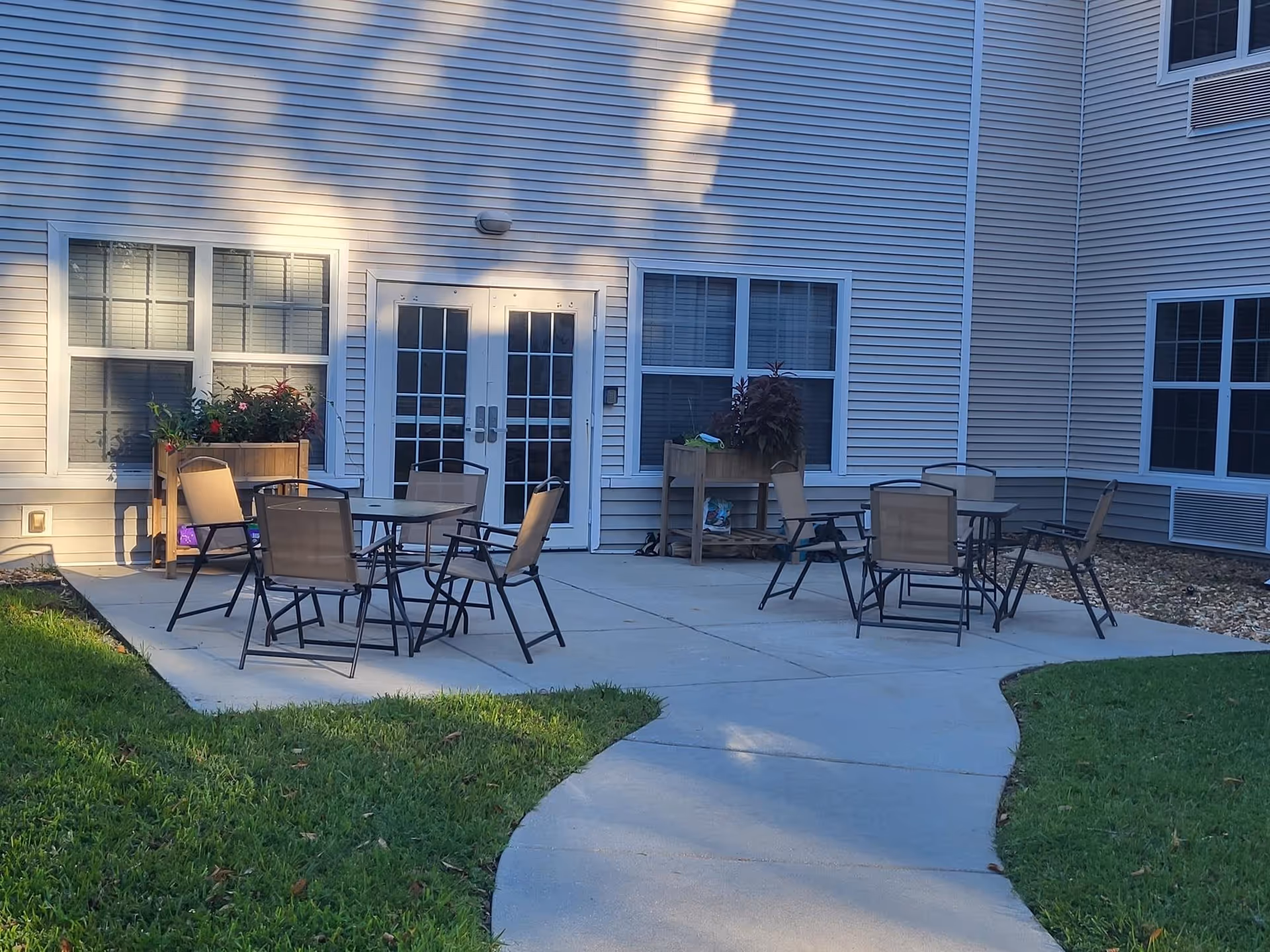 Outdoor patio area with two sets of glass-top tables and several beige mesh chairs on a concrete surface. The patio is adjacent to a building with white siding, multiple windows, and a set of double glass doors. There are two wooden planters with flowers and plants near the building. Green grass surrounds the patio area.