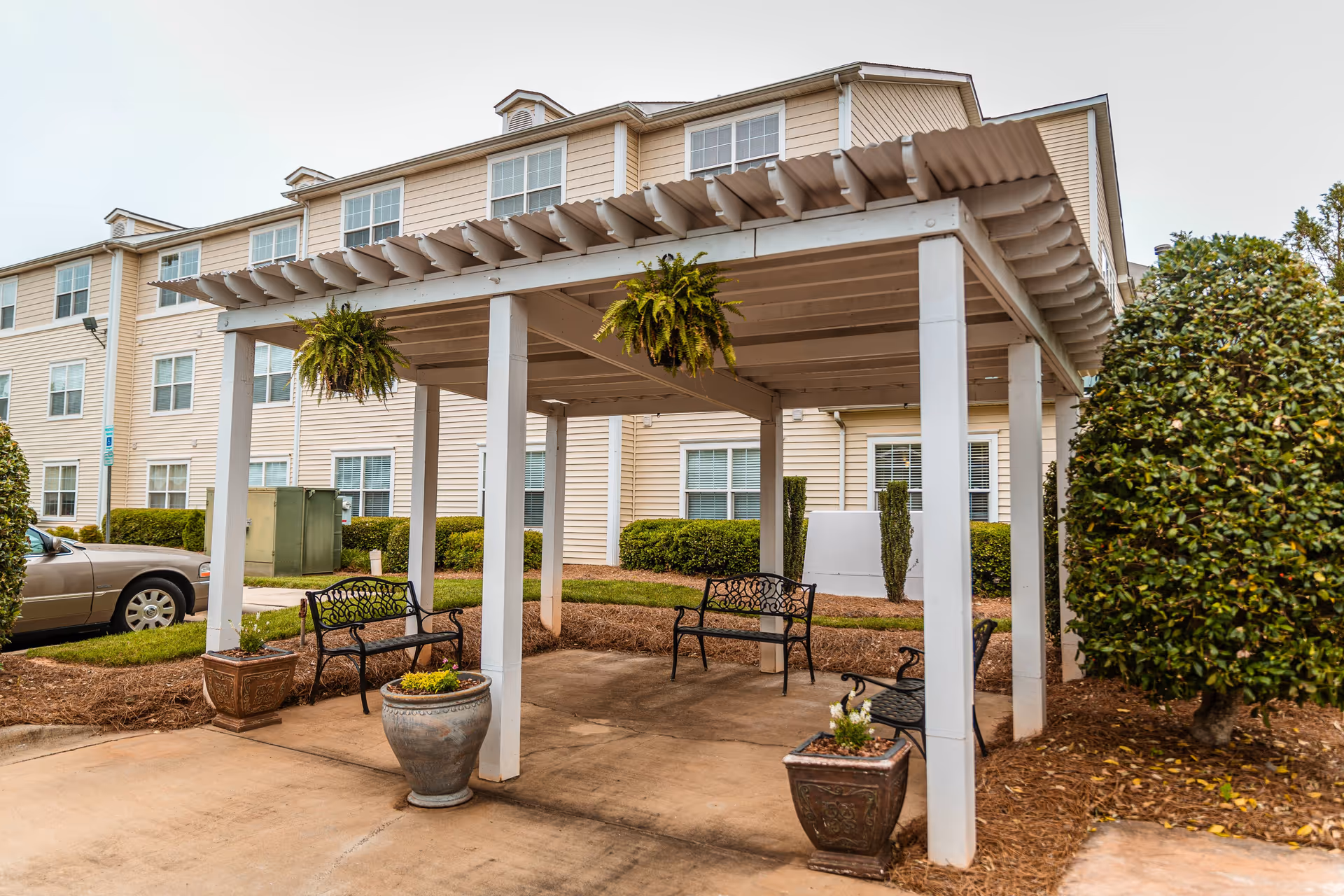 Outdoor seating area with a white pergola structure, black metal benches, hanging plants, and potted plants in front of a beige multi-story building with multiple windows.