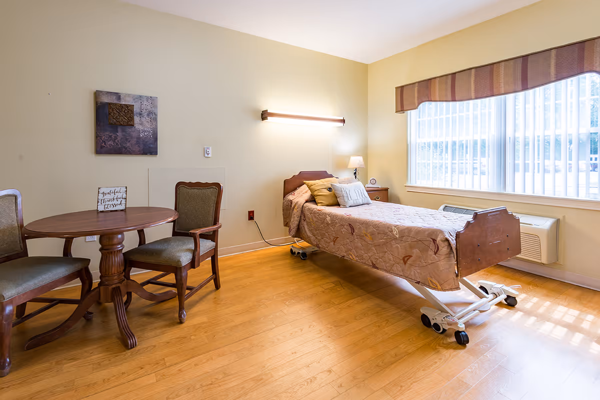 A bright senior living bedroom with a single adjustable bed covered in a patterned quilt, two pillows, a bedside table with a lamp and clock, a round wooden table with two upholstered chairs, a large window with vertical blinds and a valance, and a piece of wall art above the table.