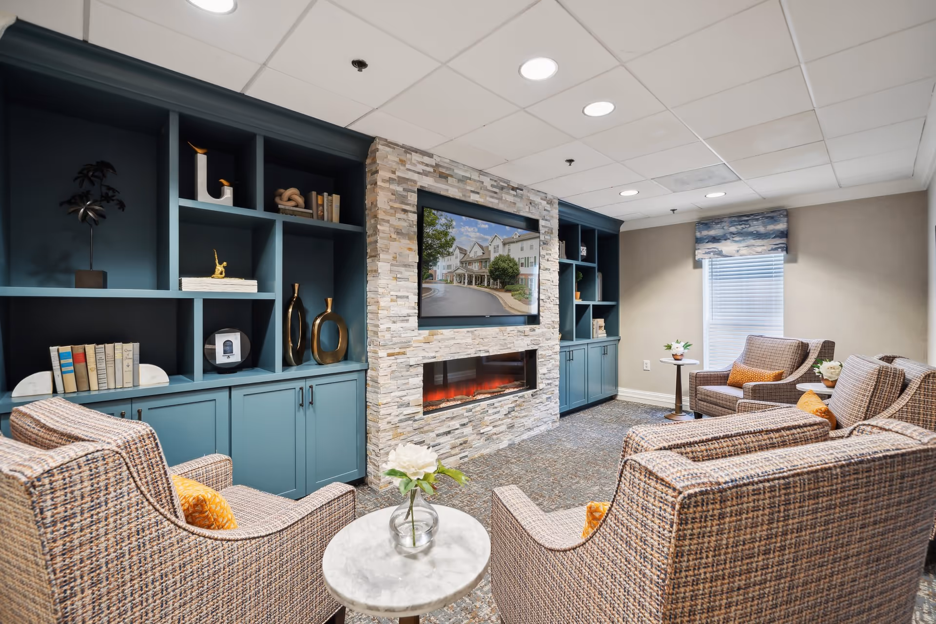 Cozy seating area with four upholstered chairs around a small table facing a stone fireplace and wall-mounted TV surrounded by teal built-in shelves.