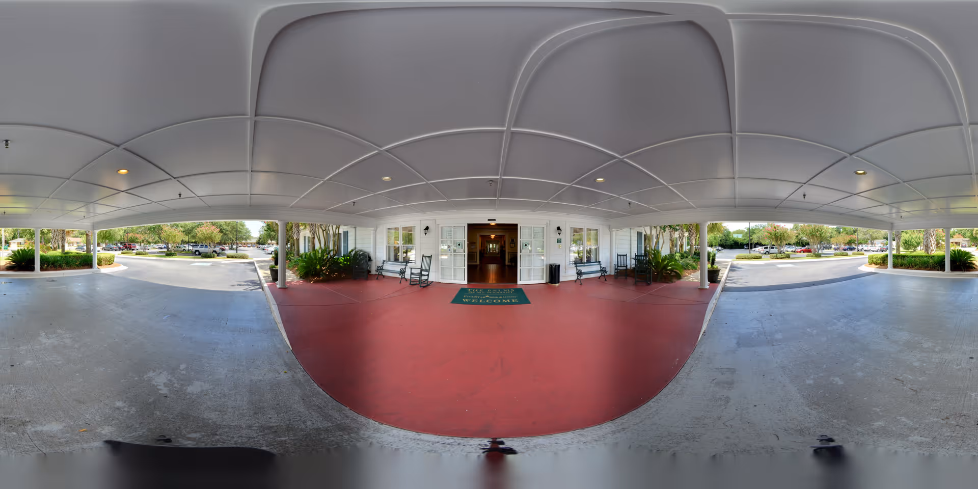 Covered entrance area of The Palms of Mt. Pleasant facility with a red floor, white ceiling with recessed lights, benches on either side of the entrance door, plants near the walls, and a green welcome mat in front of the double glass doors. Outside the covered area, there is a parking lot with cars and trees in the background.