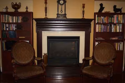 A traditional living room with a dark wood fireplace, two upholstered chairs, and bookshelves.