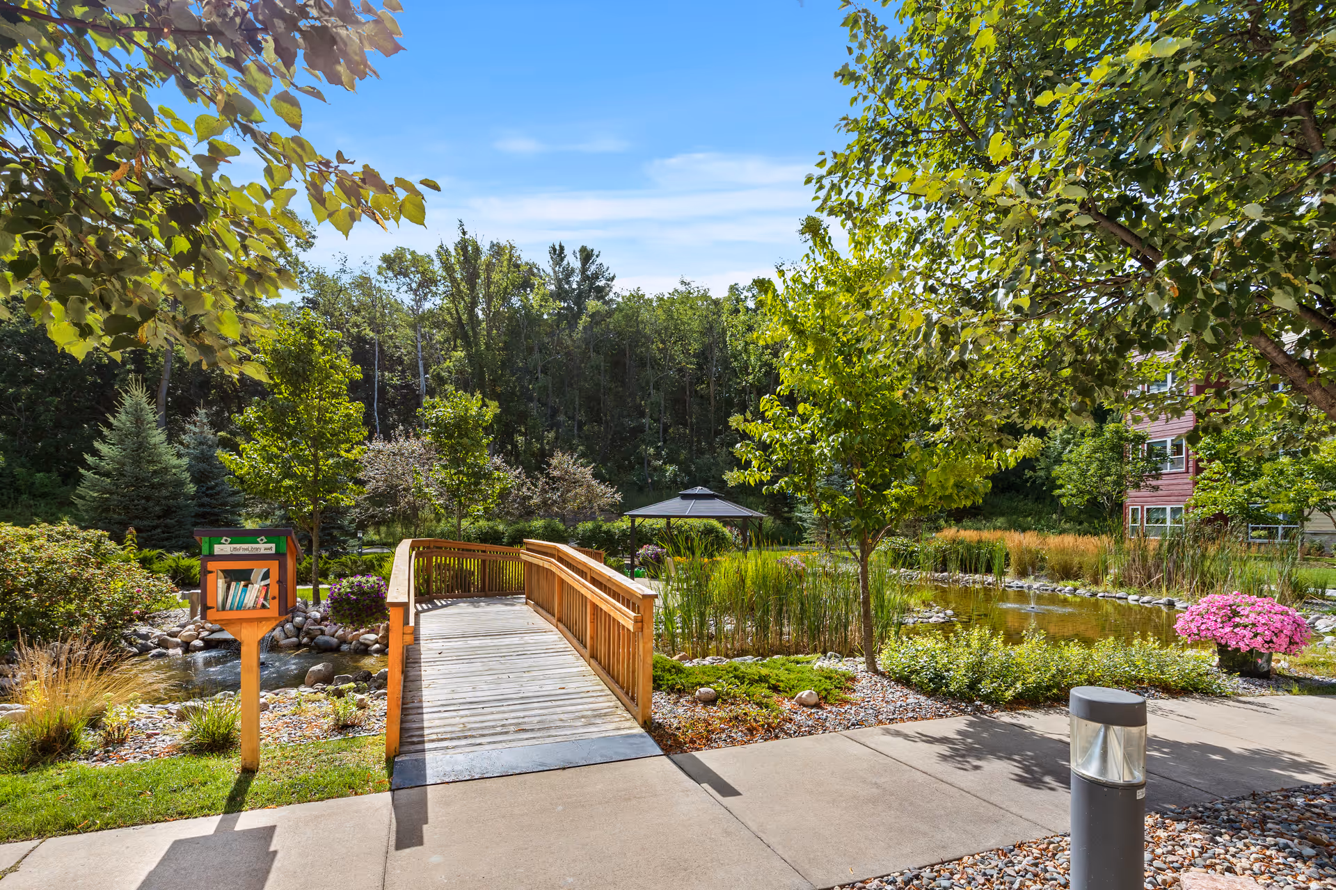 A wooden footbridge over a small pond in a landscaped garden area with trees, shrubs, and flowers. A small gazebo is visible in the background, and a little free library box with books is on the left side near the bridge. The sky is clear and blue.