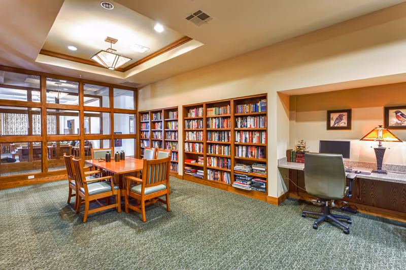 A bright senior living facility reading room with bookshelves, a wooden table and chairs, and a computer workstation.