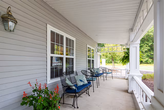 Covered porch area with gray siding walls, white framed windows, and black metal chairs with blue cushions and decorative pillows. There is a hanging plant and a lantern-style light fixture on the wall. The porch overlooks a green outdoor area with trees and a gravel path.