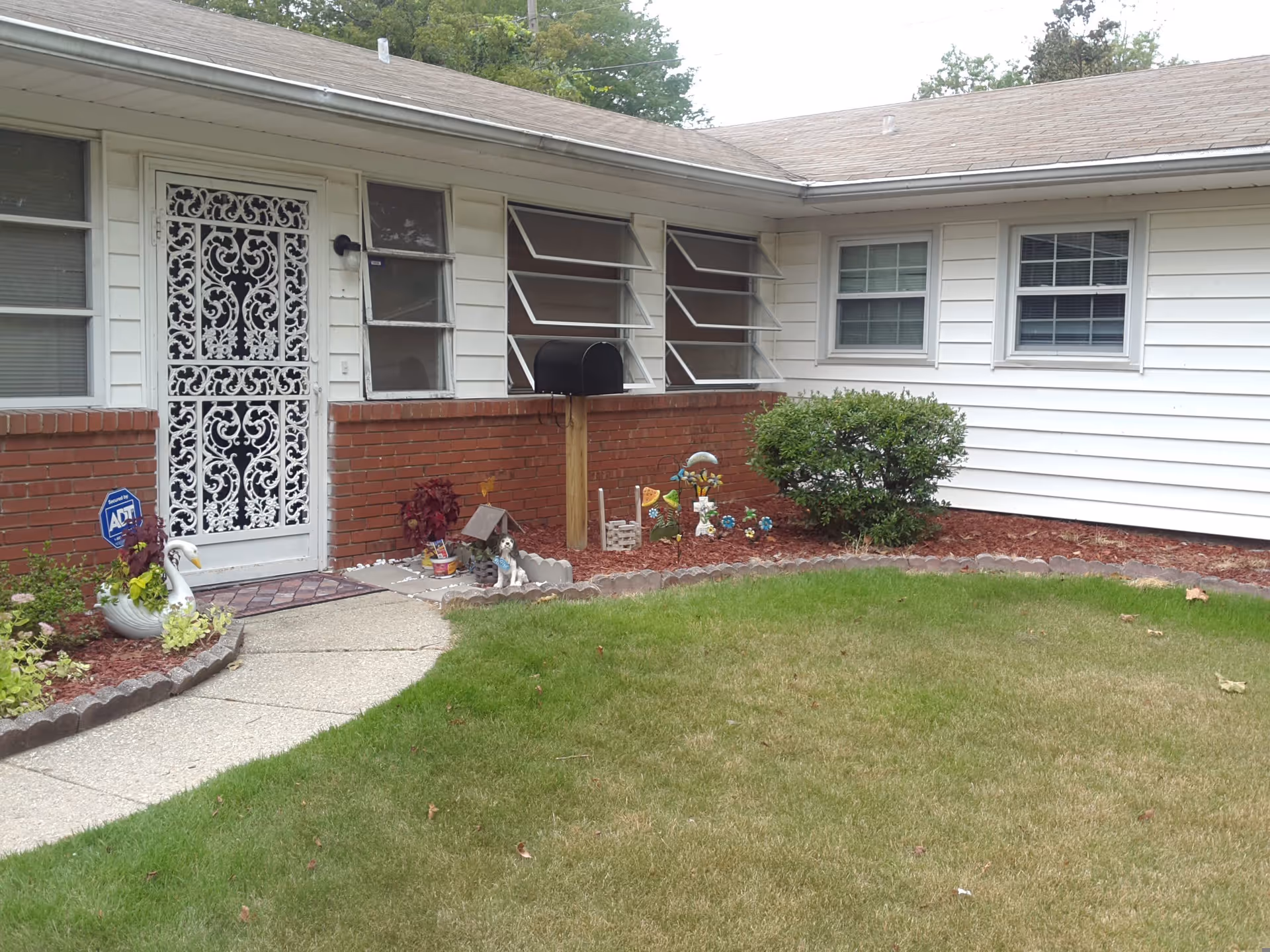 Front exterior view of a single-story building with white siding and a brick lower wall. There is a decorative white metal door, several windows, a black mailbox on a wooden post, and a small garden area with plants, a swan planter, and garden decorations. A concrete pathway leads to the door, and there is a green lawn in the foreground.