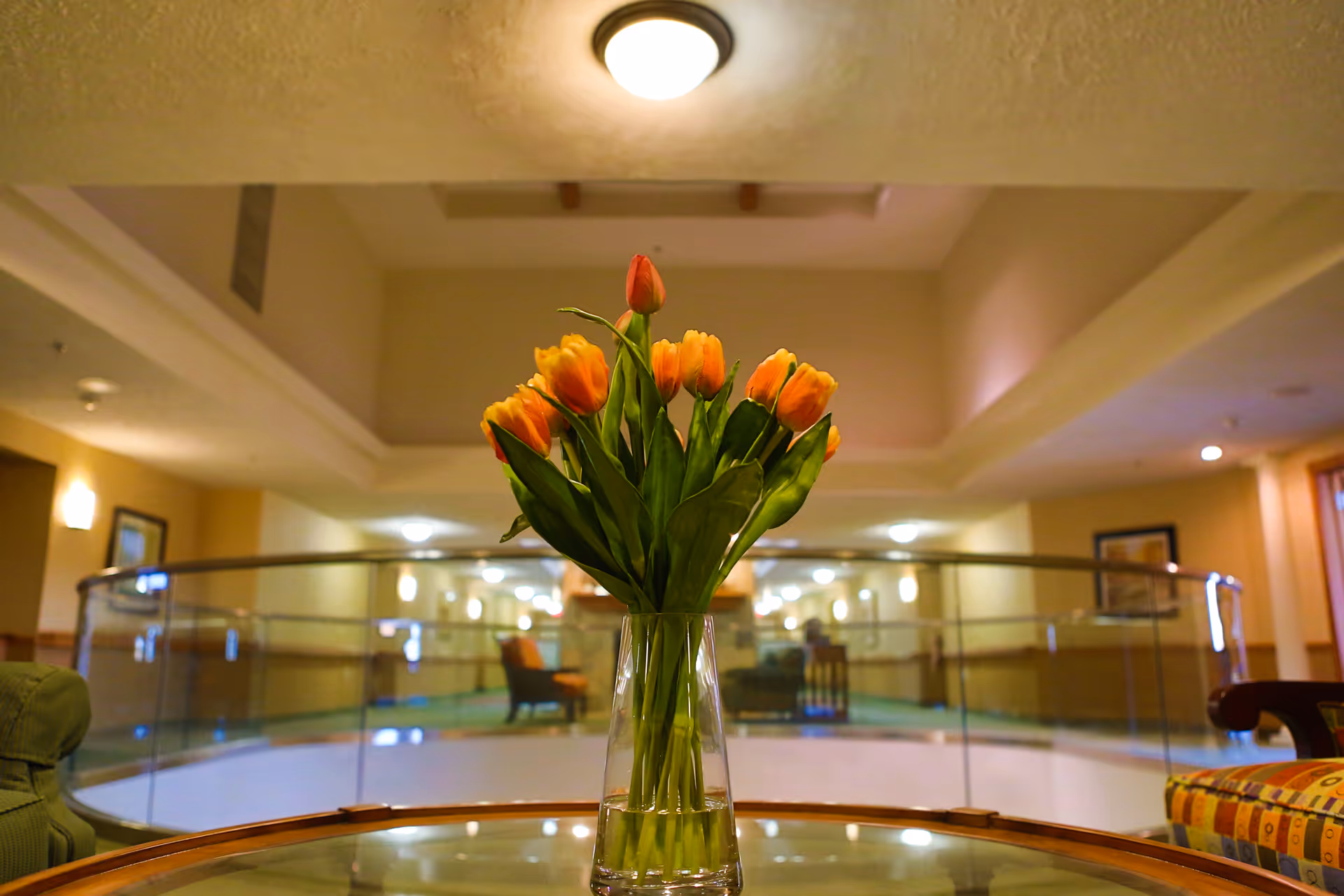 A vase of orange tulips on a glass table in a well-lit lobby/common area with seating and a curved glass railing in the background.