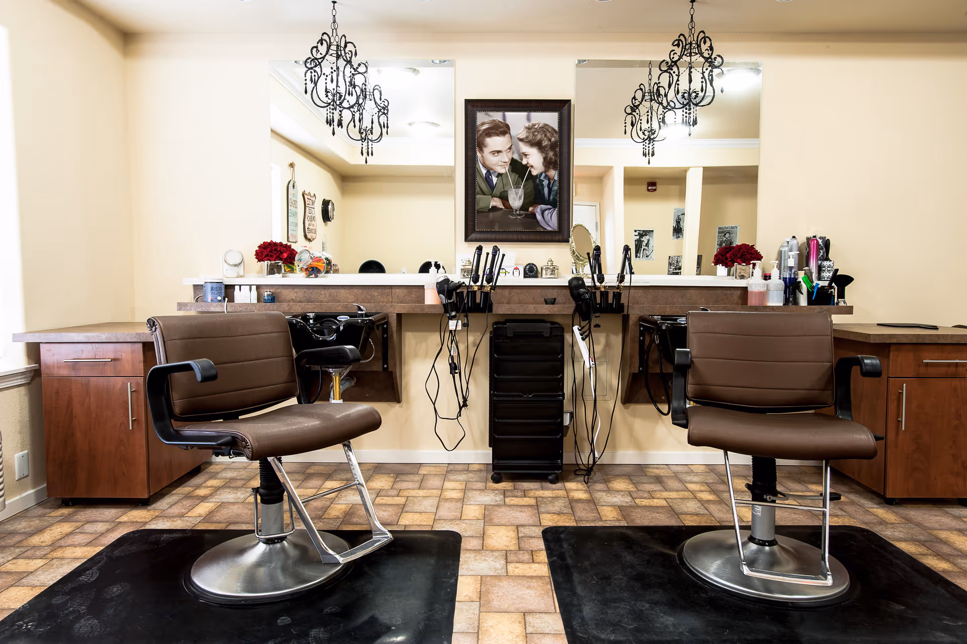 Interior view of a hair salon area with two brown salon chairs facing large mirrors. The space includes hair styling tools, a black rolling cart, and decorative elements such as a framed vintage photo of a couple sharing a milkshake and two chandeliers hanging from the ceiling.