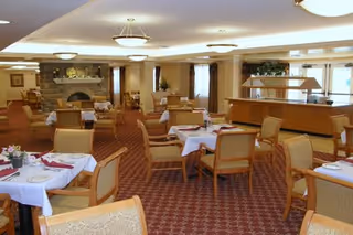 Dining room with neatly set tables and chairs, a buffet station, and a fireplace in a senior living facility.