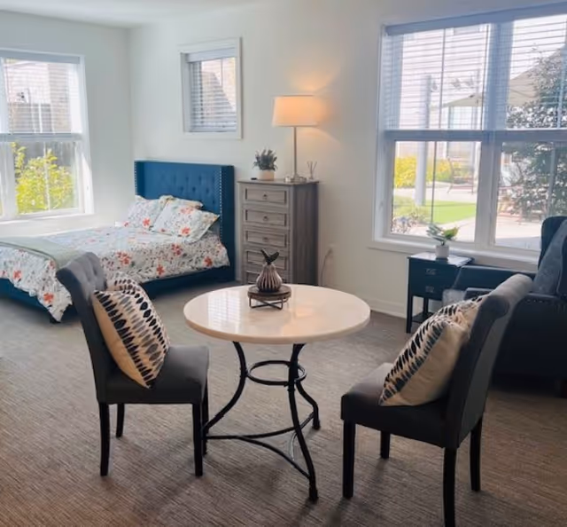 A bright studio bedroom with a floral bed, a round table with two upholstered chairs, a dresser and large windows.