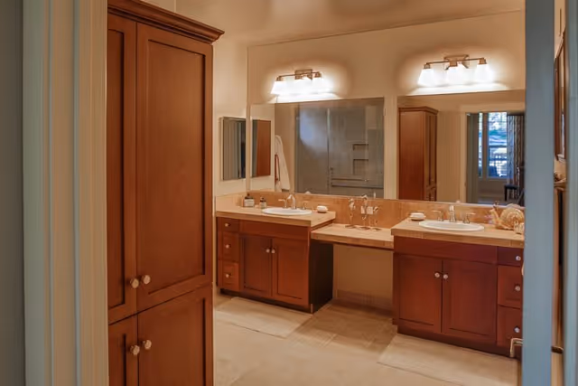 A bathroom with two sinks set in a long countertop with wooden cabinets underneath. Above the sinks is a large mirror with two light fixtures mounted on the wall. To the left, there is a tall wooden cabinet. The floor is tiled, and a shower area is visible in the reflection of the mirror.
