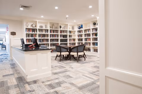 A bright and spacious library room with white built-in bookshelves filled with books and decorative items. In the center, there is a round wooden table surrounded by four cushioned chairs. The floor is covered with a patterned carpet in neutral tones, and the ceiling has recessed lighting. A reception desk with a computer is visible on the left side near the entrance.
