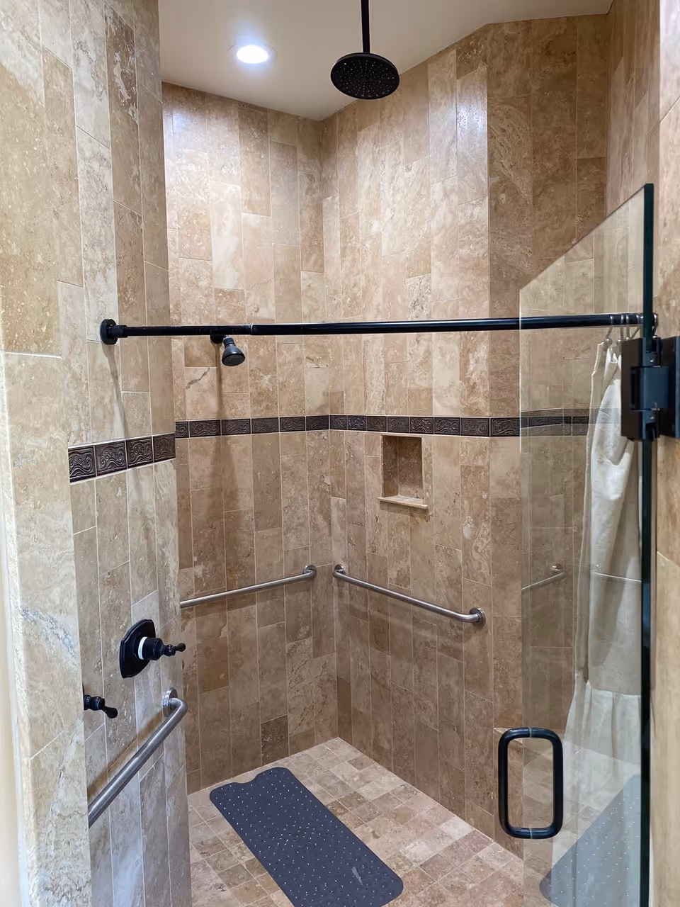 A walk-in shower with beige marble tiles covering the walls and floor. The shower features a black overhead rain showerhead, a smaller adjustable showerhead, and multiple stainless steel grab bars for safety. There is a built-in niche for toiletries and a black non-slip mat on the floor. The shower is enclosed with a glass door with a black handle.