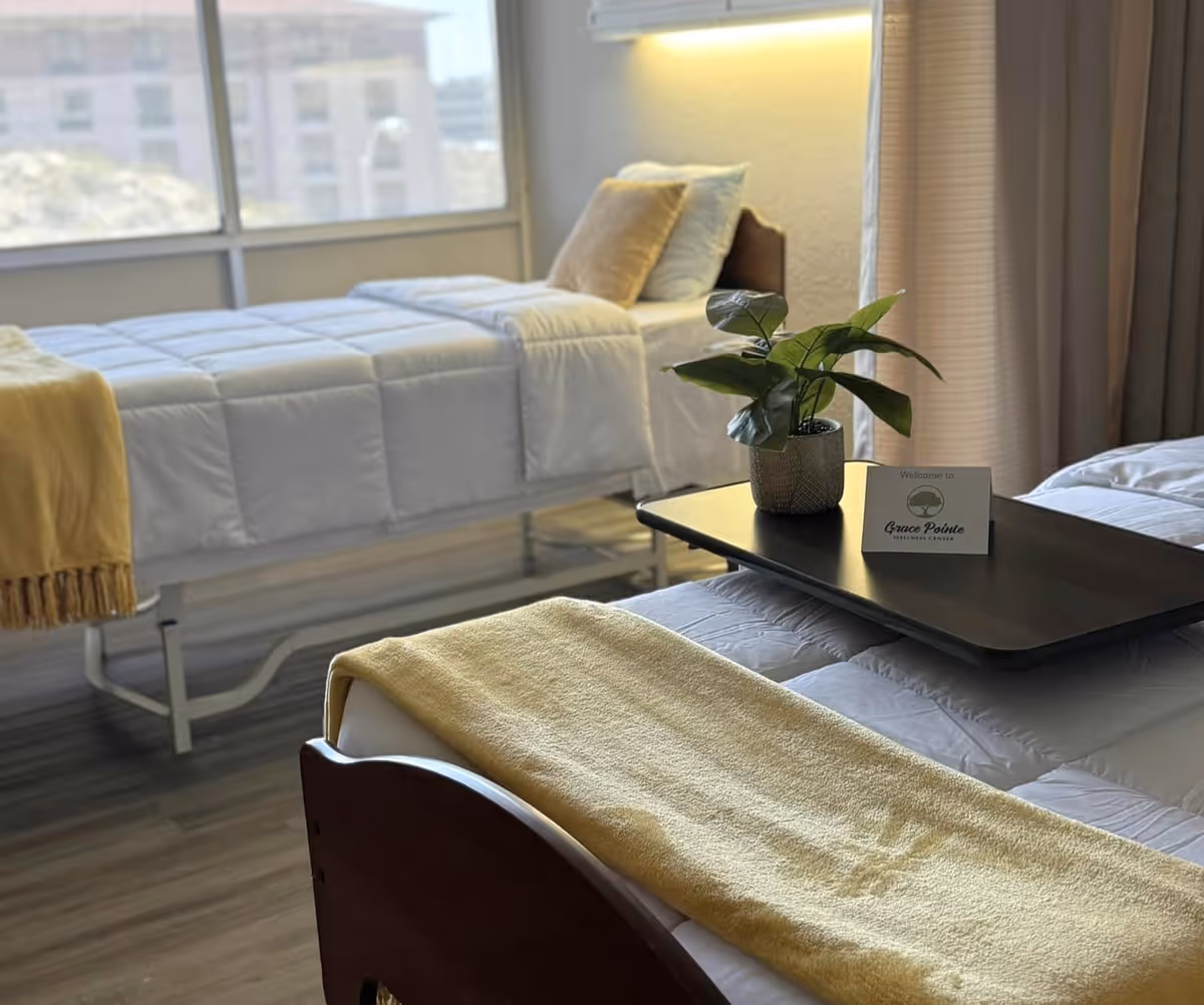 A well-lit bedroom with two single beds, each made with white bedding and a yellow blanket at the foot. A small black tray table on one bed holds a potted plant and a card that reads 'Welcome to Grace Pointe Wellness Center.' Large windows in the background let in natural light.
