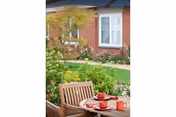Outdoor patio area with a wooden table and chairs set with red cups and plates, surrounded by green plants and flowers, with a brick building and windows in the background.