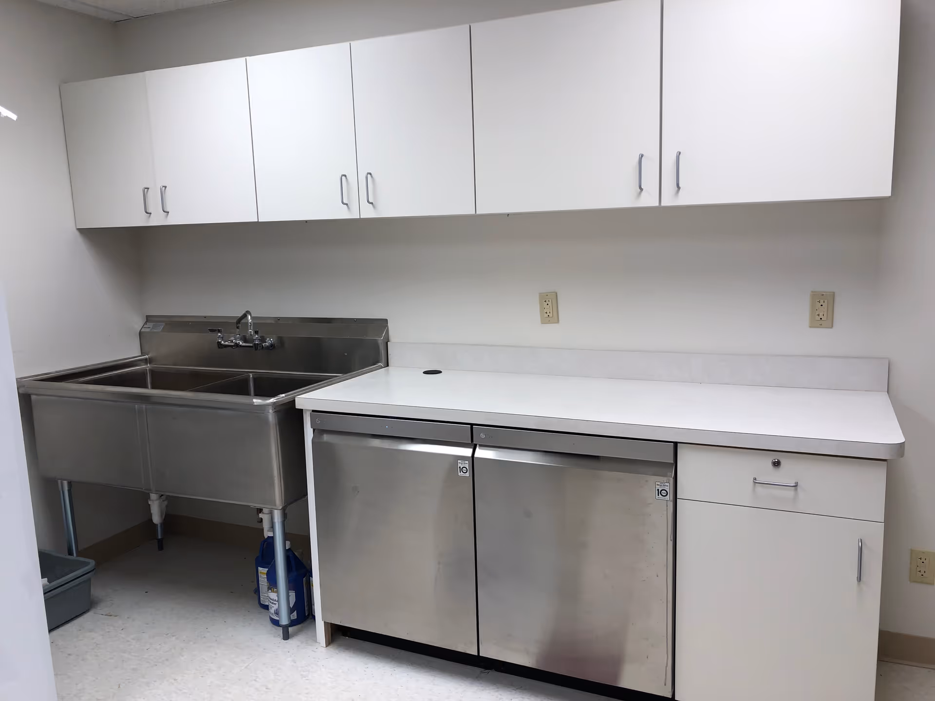 A clean utility or kitchen area with a large stainless steel double sink on the left, white cabinets above, and a white countertop with stainless steel under-counter refrigerators or storage units below. The walls and floor are plain and light-colored.