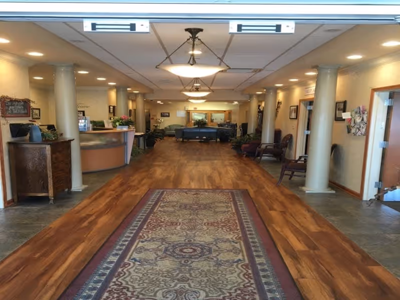 Interior view of a senior living facility hallway with wooden flooring and a patterned rug in the center. The hallway is lined with large white columns on both sides, with chairs and small tables along the walls. Ceiling lights illuminate the space, and a reception desk is visible on the left side near the entrance. The far end of the hallway has seating areas with couches and plants.