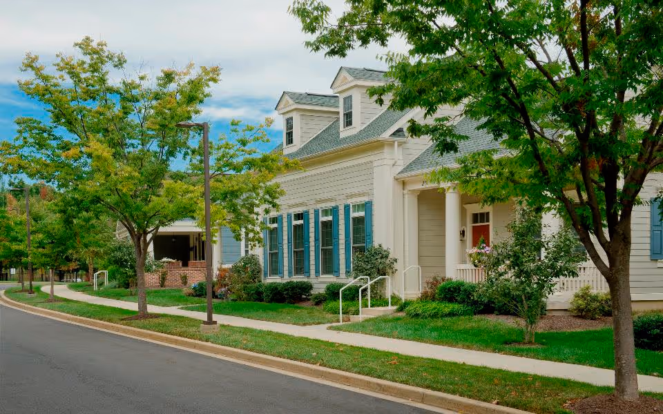 Exterior view of a residential-style building with beige siding, blue shutters, and a green roof. The building is surrounded by green trees and shrubs, with a sidewalk and street in the foreground under a partly cloudy sky.