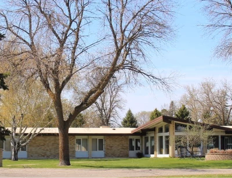 Single-story building with large windows and a slanted roof surrounded by leafless trees and green grass under a clear blue sky.