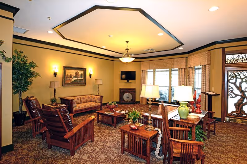 A warmly lit living room with wooden furniture including armchairs, a sofa, coffee tables, and side tables with lamps. The room features a patterned carpet, a large window with curtains, a wall-mounted TV, a framed painting, and decorative plants. The entrance door has an artistic tree design.