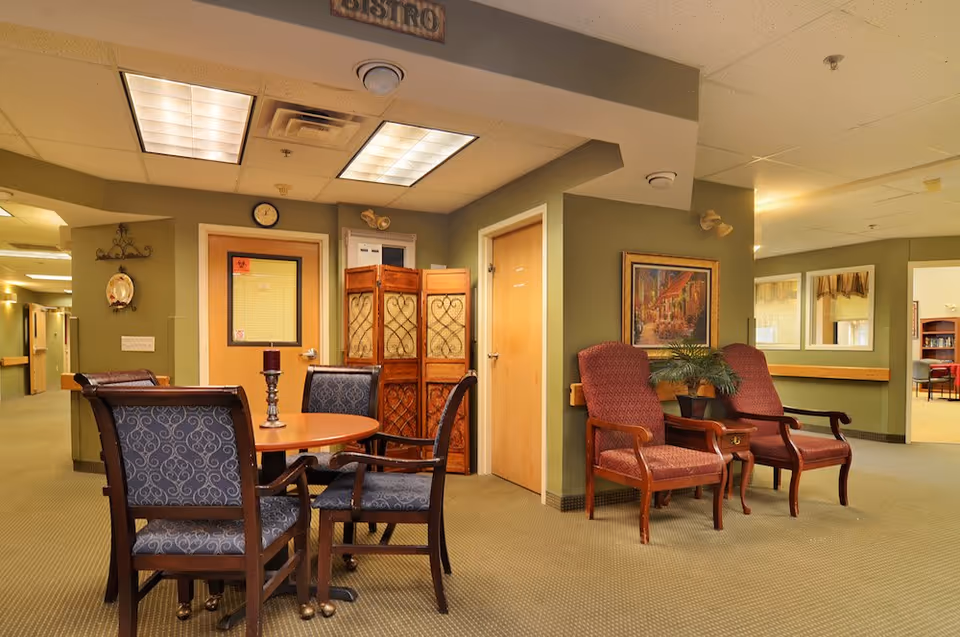 Interior view of a senior living facility common area with a round wooden table surrounded by four upholstered chairs, two red cushioned armchairs with wooden arms, a small side table with a potted plant, a decorative folding screen, and a framed painting on the wall. The walls are painted green and the ceiling has recessed lighting panels. A sign above reads 'Bistro'.