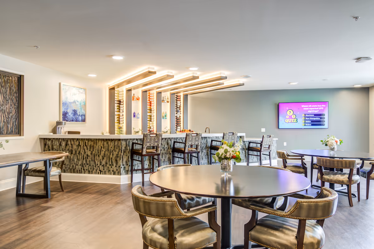 A modern common area with a bar counter featuring a decorative tile front and illuminated overhead beams holding wine bottles. There are several bar stools at the counter and round tables with cushioned chairs arranged around the room. A flat-screen TV on the wall displays a quiz question about UFO sightings. The room has wood flooring and light-colored walls with artwork and floral centerpieces on the tables.