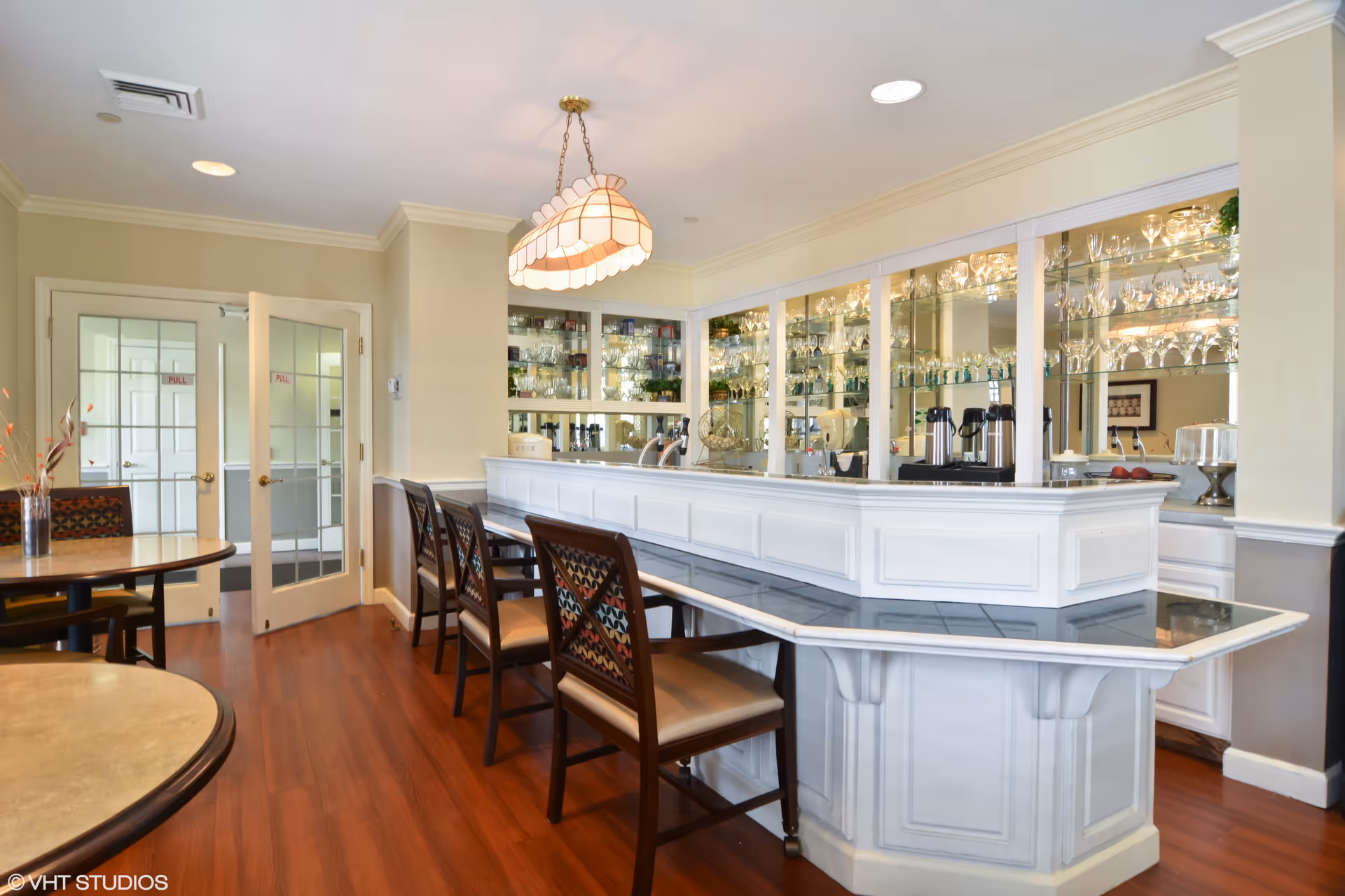 Interior view of a dining area with a white bar counter and several wooden chairs with patterned cushions. Behind the bar is a mirrored wall with shelves displaying various glassware. There are round tables with chairs in the foreground and double glass doors in the background. The floor is wooden, and a decorative hanging light fixture is above the bar.