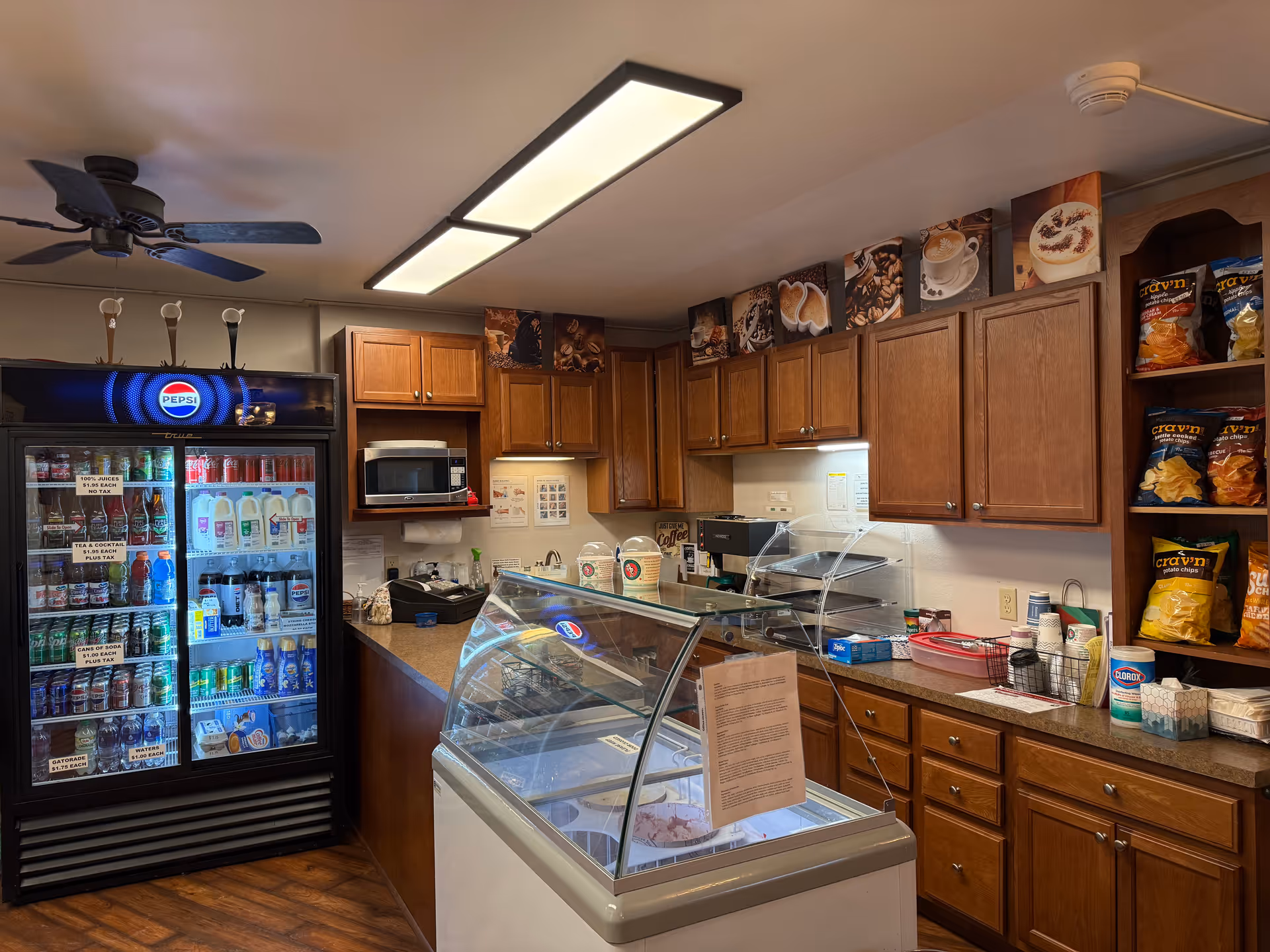 Communal snack counter with a glass-front beverage refrigerator, curved ice cream display, and wooden cabinets stocked with chips and supplies.