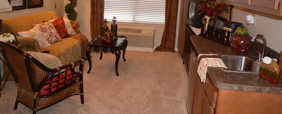 Cozy studio living area with a sofa and wicker chair around a coffee table, a window with blinds, and a small kitchenette with sink and countertop.