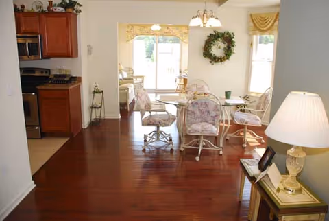 Open interior showing a dining area with floral-upholstered chairs around a glass table on polished hardwood floors, with a kitchen to the left and a living area beyond.