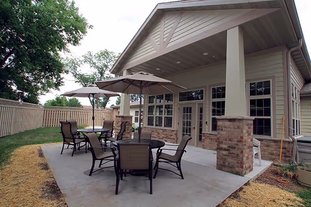 Outdoor patio area at a senior living facility with multiple round tables and chairs under large umbrellas, adjacent to a building with large windows and brick pillars, surrounded by a wooden fence and trees.