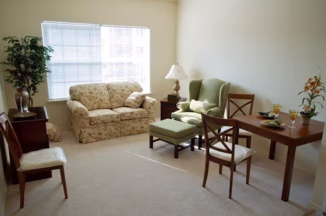 Bright furnished living room with a floral loveseat, green wingback chair and ottoman, a small wooden dining table with chairs, and a window.