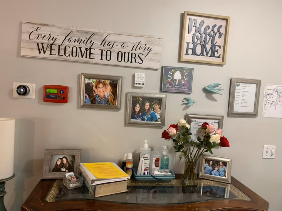 A wall decorated with framed photos, inspirational signs, and two decorative blue birds. The signs include messages such as 'Every family has a story WELCOME TO OURS' and 'Bless our HOME'. Below the wall is a wooden table with a glass top holding a vase of red, pink, and white roses, hand sanitizer bottles, a binder, and a small framed photo. A thermostat and fire alarm are mounted on the wall.