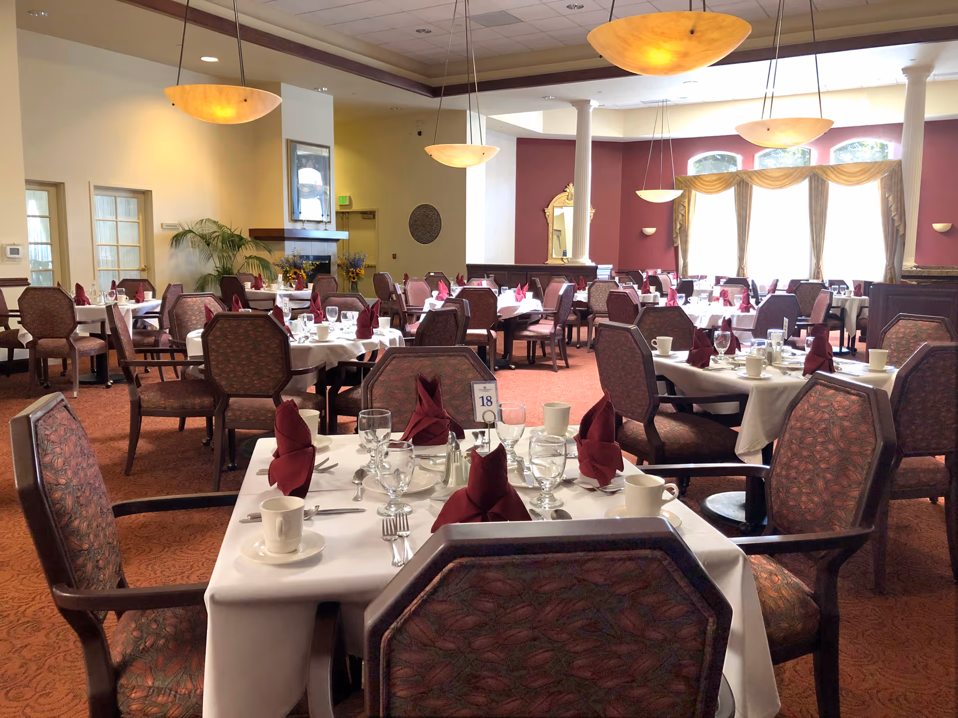 A spacious dining room with multiple tables covered in white tablecloths, each set with glassware, cups, silverware, and folded burgundy napkins. The room features patterned upholstered chairs, warm lighting from hanging pendant lamps, large windows with gold curtains, and a fireplace in the background.