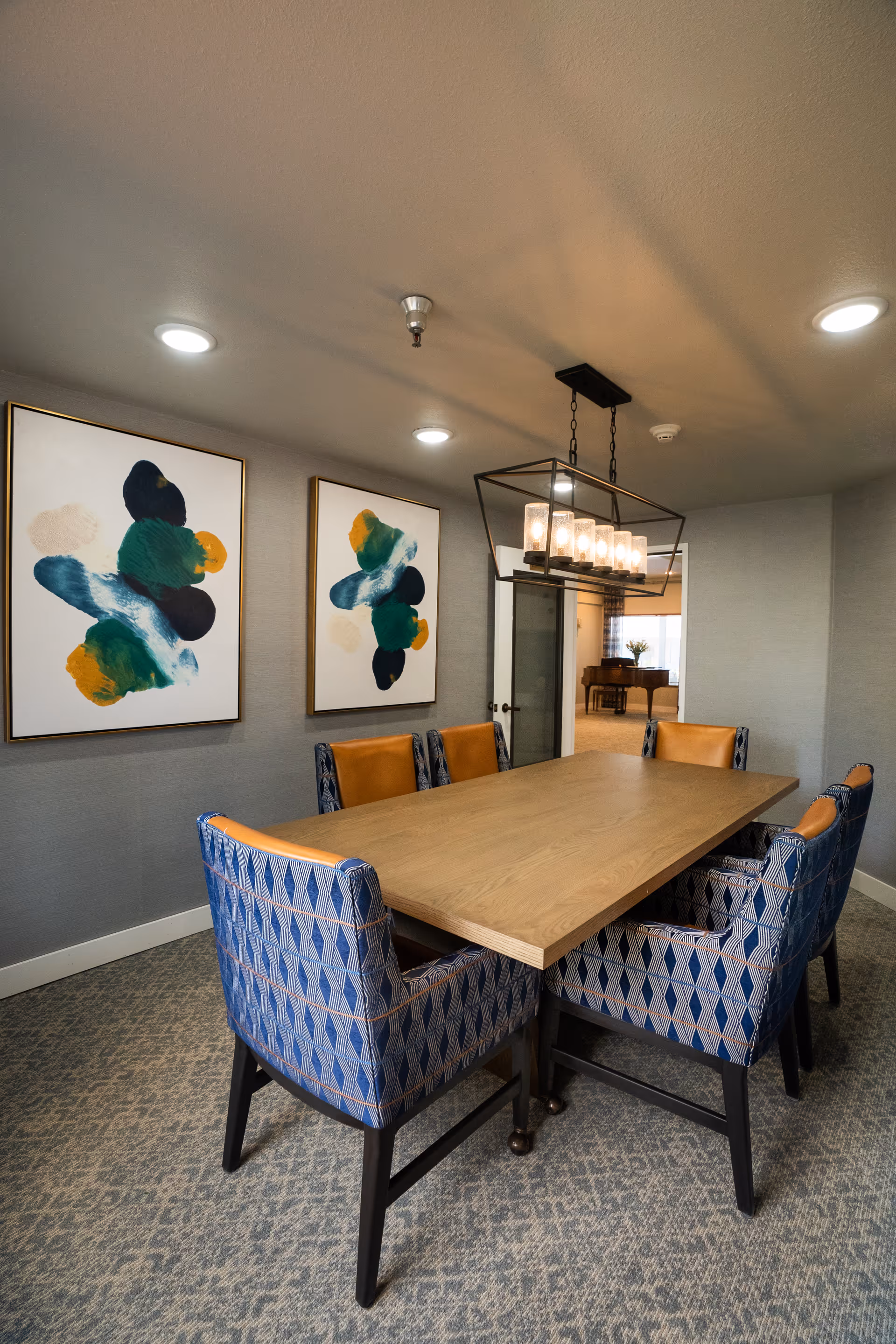 Rectangular wooden dining table surrounded by patterned upholstered chairs beneath a rectangular chandelier, with abstract paintings on the wall.