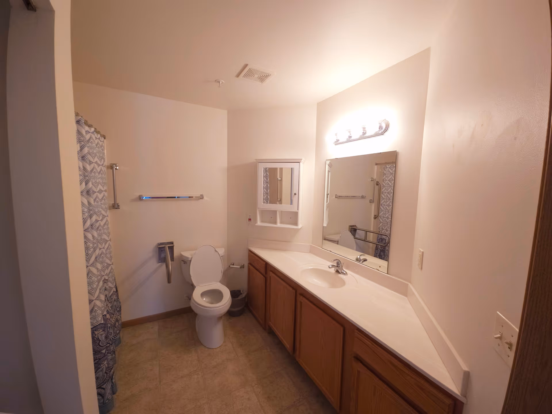A bathroom with a toilet, a shower with a patterned curtain, a long countertop with a sink, a large mirror above the sink, wooden cabinets below the countertop, and a wall-mounted cabinet with a mirror. The walls are light-colored and the floor is tiled.