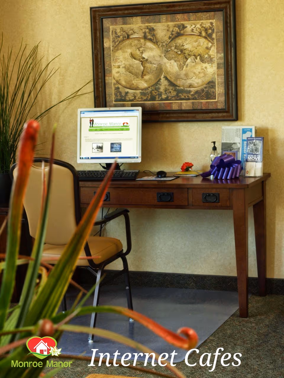 A small interior internet café area with a computer on a wooden desk, a chair, wall art, and plants in the foreground.