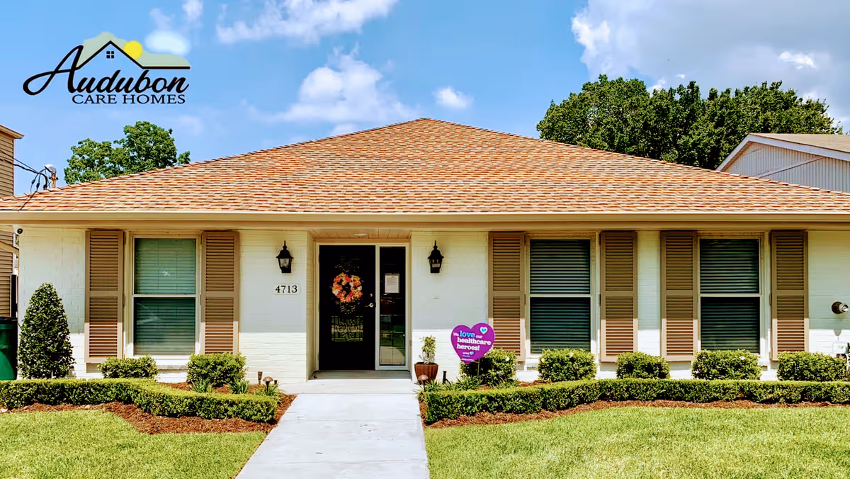 Front exterior view of a single-story building with a tan shingled roof, white brick walls, and brown shutters on the windows. A concrete walkway leads to a glass front door decorated with a wreath. There are two black wall-mounted lanterns on either side of the door, neatly trimmed bushes along the front, and a purple heart-shaped sign that says 'We love healthcare heroes!'. The sky is blue with some clouds.