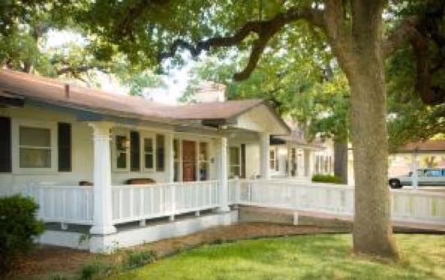 Exterior view of a single-story assisted living facility building with a white wooden porch railing, several windows, and a large tree in the foreground providing shade.